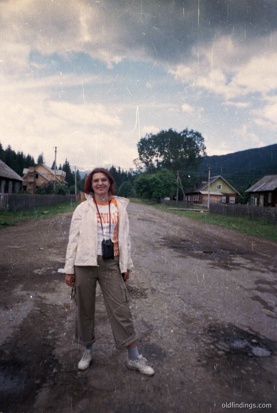 Vintage photograph of a woman in a rural village setting, likely Eastern Europe, 1970s–1980s. She wears a light jacket, patterned scarf, and beige pants with white sneakers, posing on a muddy road. Wooden houses with pitched roofs and lush green hills in the background. Camera strap suggests travel or photography interest.