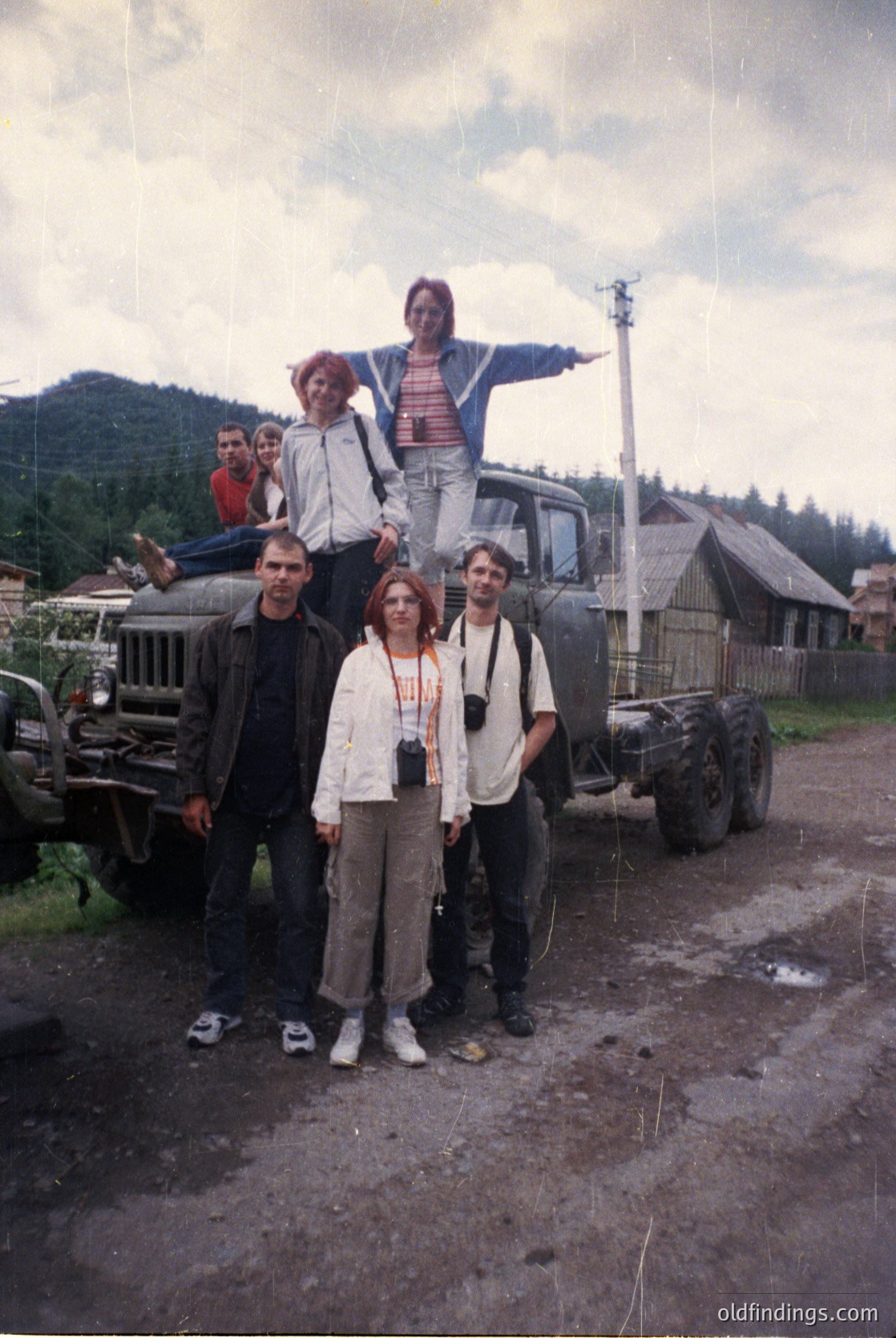 Vintage group photo on a rural dirt road, featuring six individuals posing with a Soviet-era GAZ truck. Three adults stand front-row, while three children perch atop the vehicle. Wooden houses and forested hills in the background suggest a countryside setting, likely Eastern Europe, 1960s–1980s. Casual attire and the truck’s design indicate mid-century rural life.