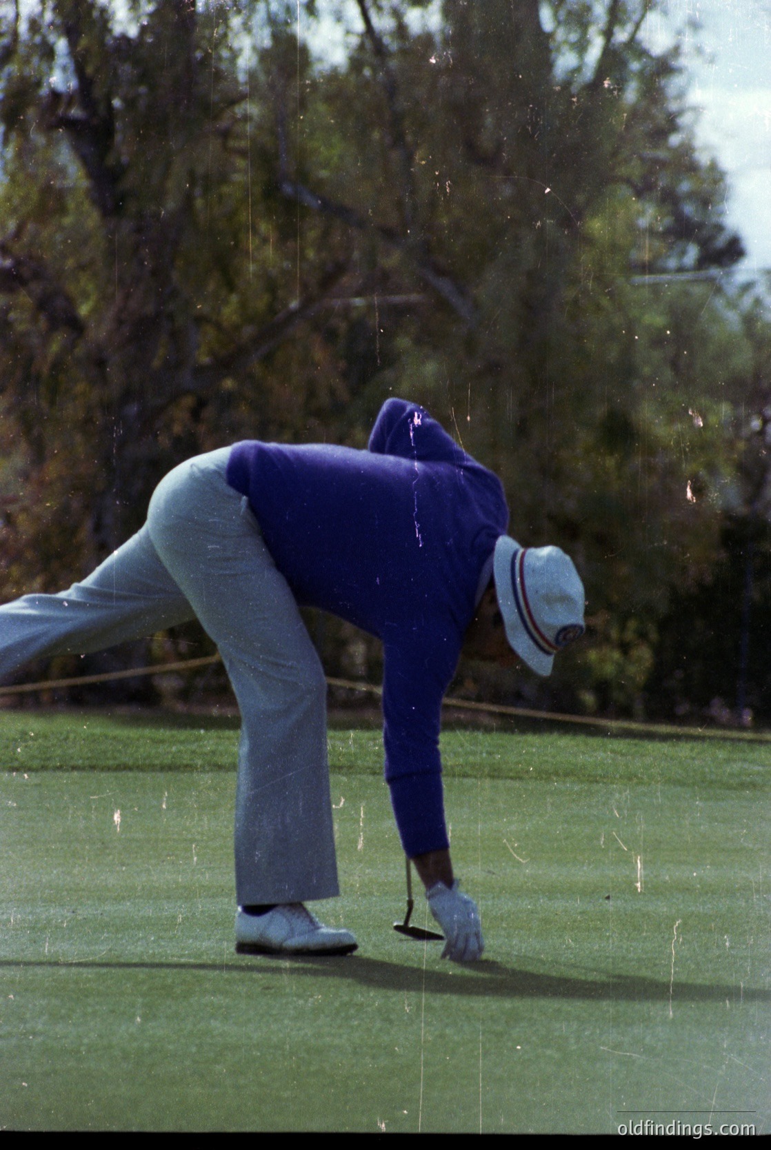 Mid-20th-century golfer in classic attire—light gray trousers, blue sweater, and white cap—executing a putt on a lush green course. Raindrops and blurred motion suggest recent or ongoing light precipitation. Dense tree line in background indicates a wooded fairway. Ideal for vintage sports, golf history, or retro lifestyle archives.