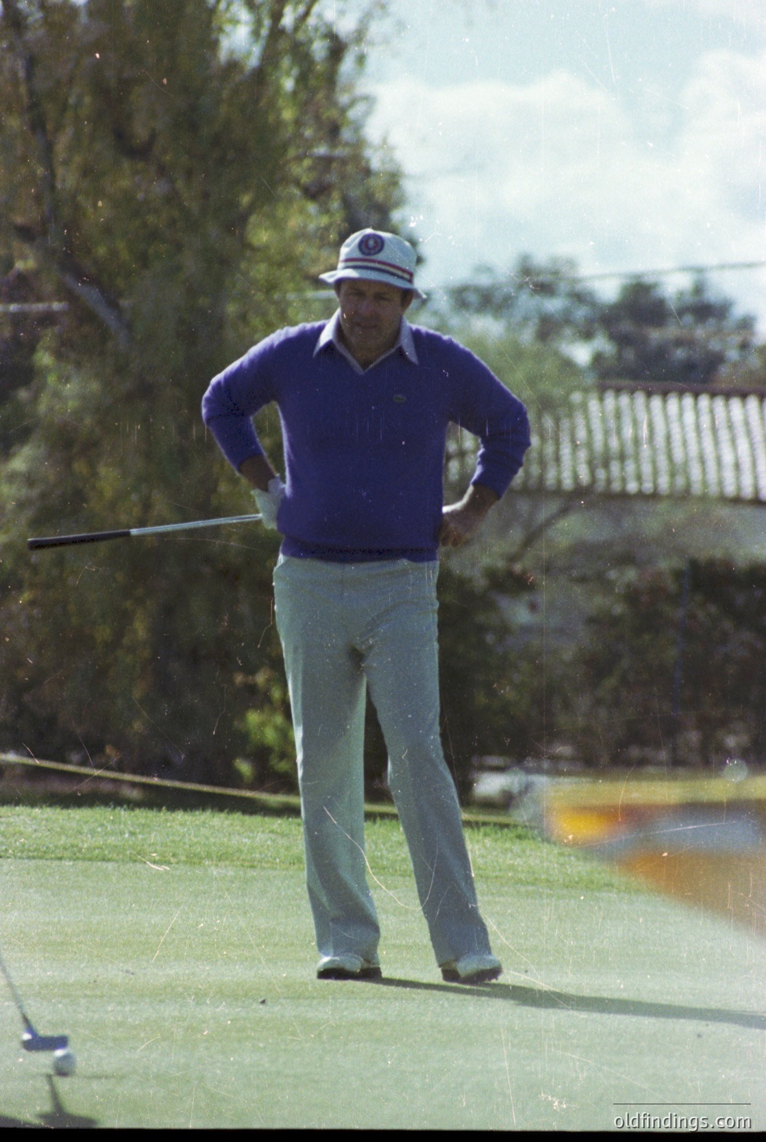 Mid-1970s golf scene: Man in vintage attire—white cap with logo, purple sweater, light trousers—mid-swing on a lush green fairway. Classic wooden club and vintage golf ball in motion. Residential backdrop with wooden fence and trees.