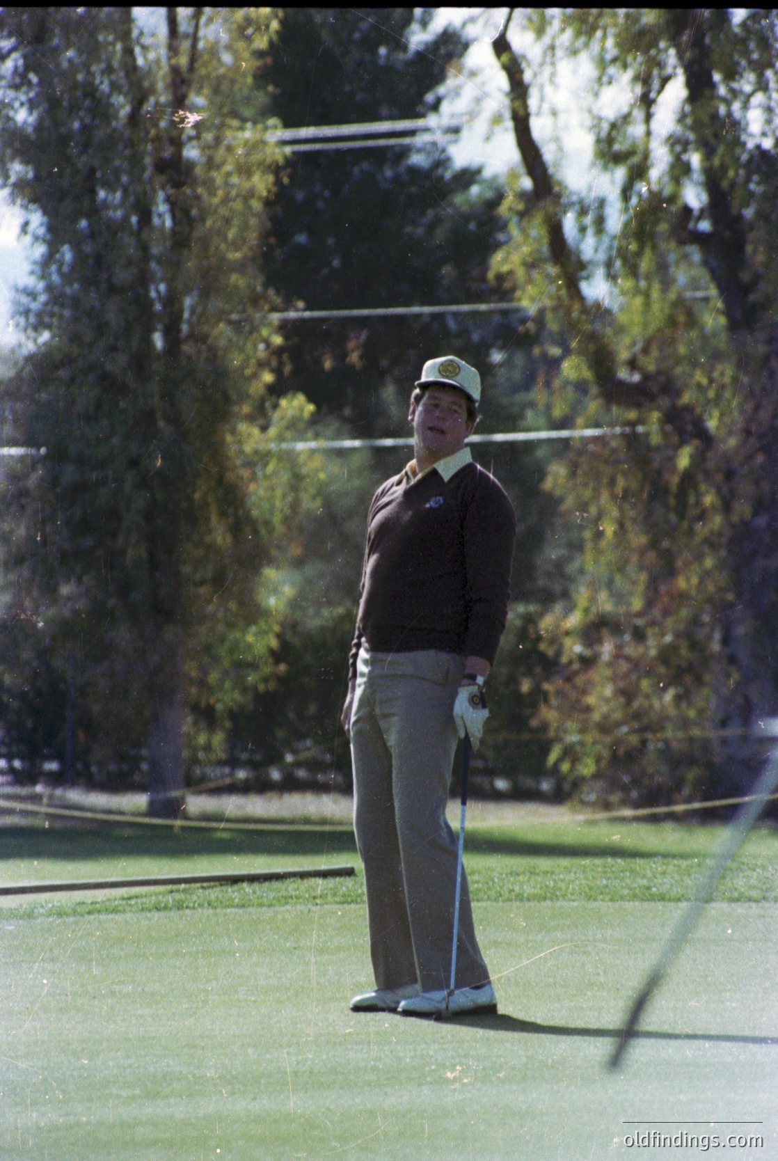 Mid-20th century golfer in vintage attire—light-colored cap, dark sweater, and slacks—poised on a lush green fairway. Golf club gripped in right hand, stance focused. Dense evergreen trees and blurred greenery in background suggest a serene, wooded course. Likely 1960s–1970s, USA or similar golf culture region.