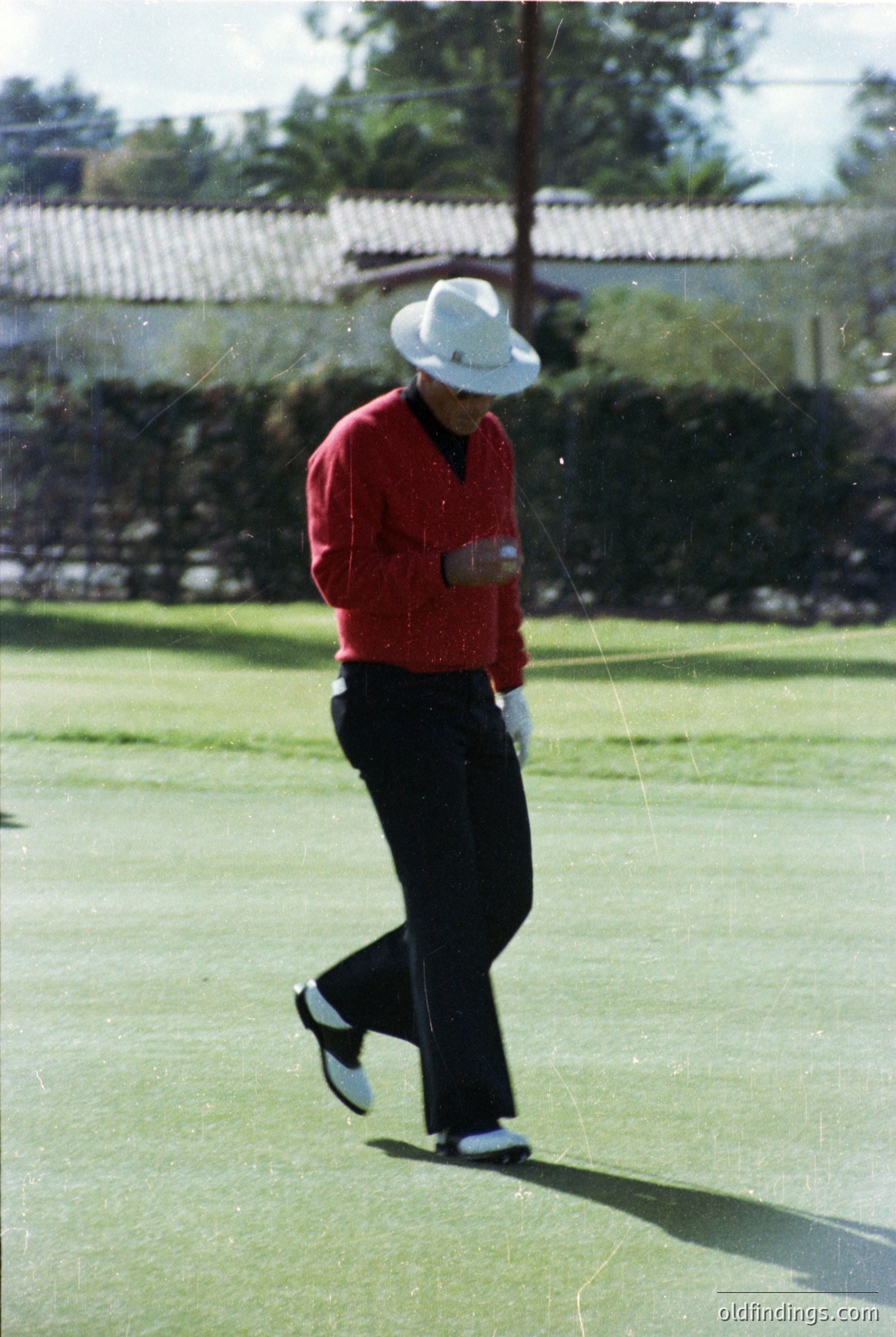 Golfer mid-swing on a manicured green, wearing a white fedora, red sweater, and black trousers with white socks and shoes. Classic 1970s golf attire. Residential-style buildings with tiled roofs in background. Ideal for vintage sports or golf history research.