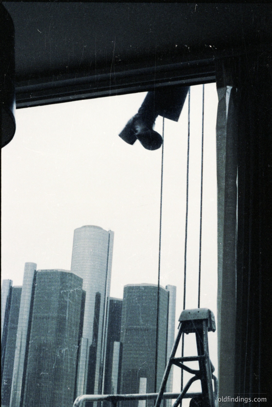 Urban skyline captured through a window with industrial lighting rigs. Mid-century modern high-rises dominate the background, suggesting a major city’s architectural style. Overhead, cables and a ladder hint at maintenance or construction activity. Likely taken in a North American metropolis during the 1960s–1980s.