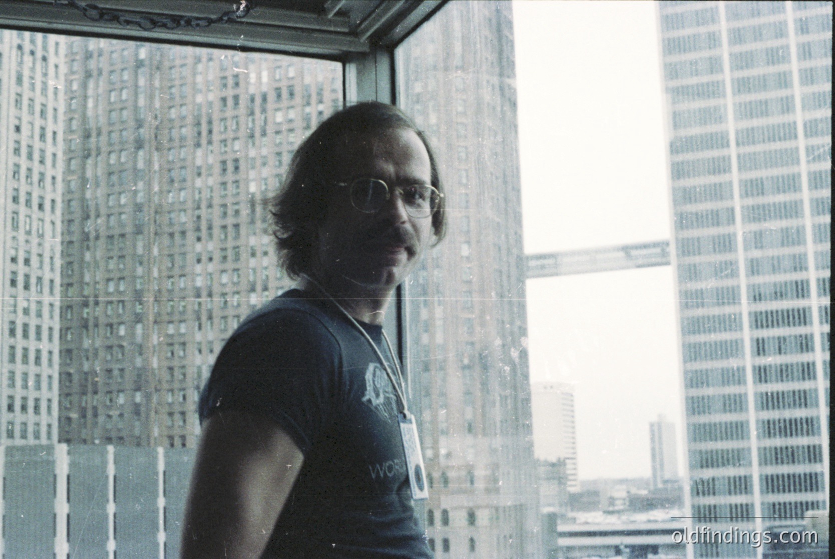 Portrait of a man in a high-rise office setting, wearing a lanyard with an ID badge reading "WORLD" and round-framed glasses. Urban skyline visible through floor-to-ceiling windows, suggesting a 1980s-1990s corporate environment.