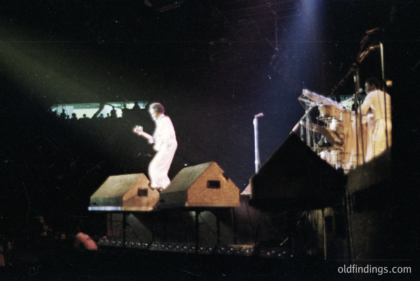 Stage performance featuring a performer in white attire atop a set resembling miniature houses, likely a 1980s rock concert. Dramatic lighting and stage props enhance theatricality.