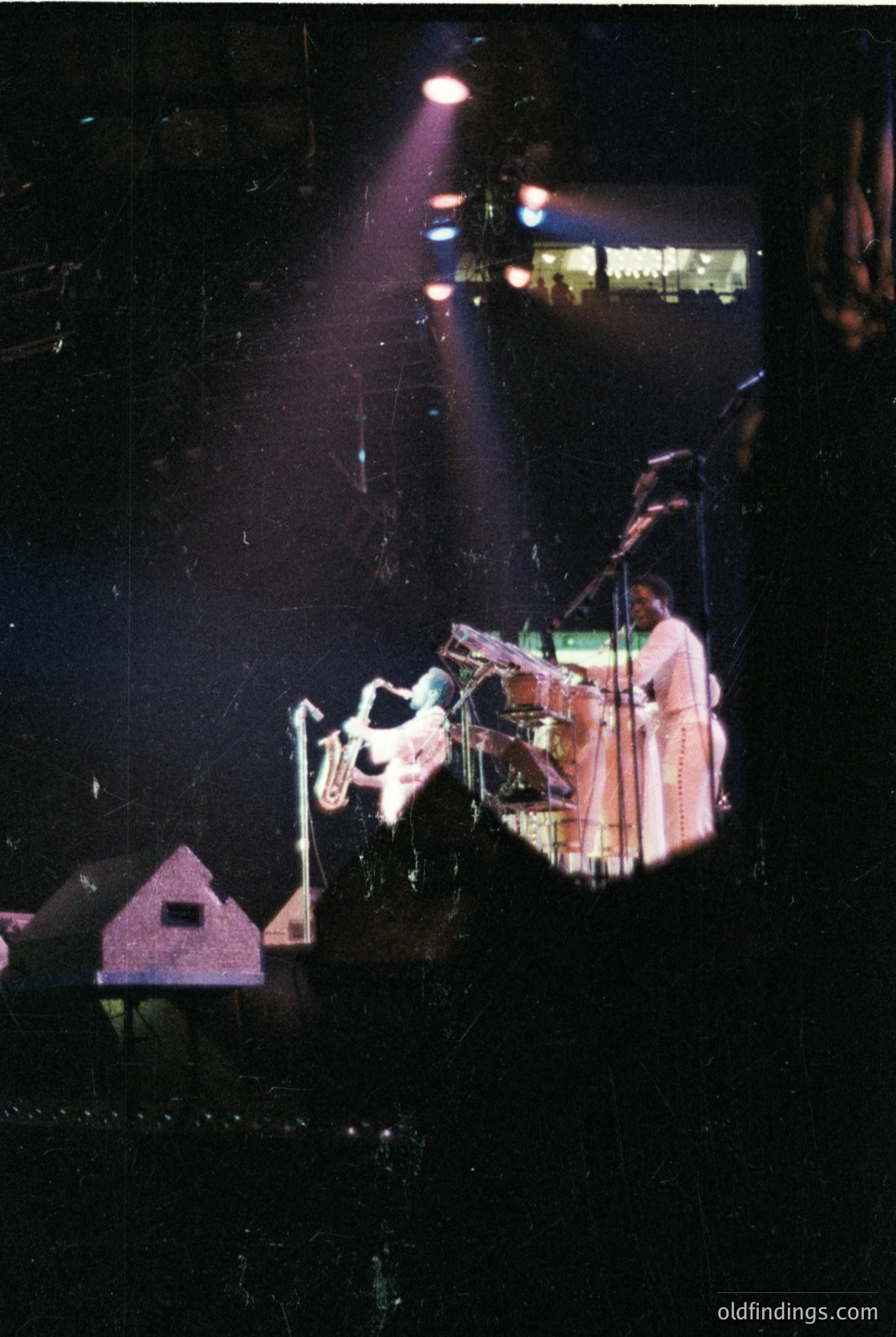 Vintage concert stage shot featuring a saxophonist in mid-performance, bathed in dramatic stage lighting. The musician wears a light-colored jacket and dark pants, playing a tenor saxophone. Backdrop includes a blurred, geometric architectural element and a small, illuminated sign. Likely from the 1960s–1970s jazz/blues era.