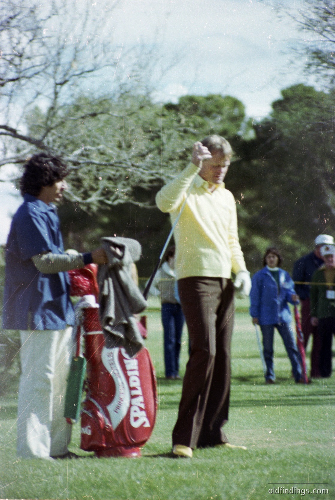 1970s golf scene featuring a golfer mid-swing in light pastel attire, red golf bag with "PGA" branding, and blurred spectators in park-like setting. Classic vintage sportswear and equipment reflect mid-century golf culture.