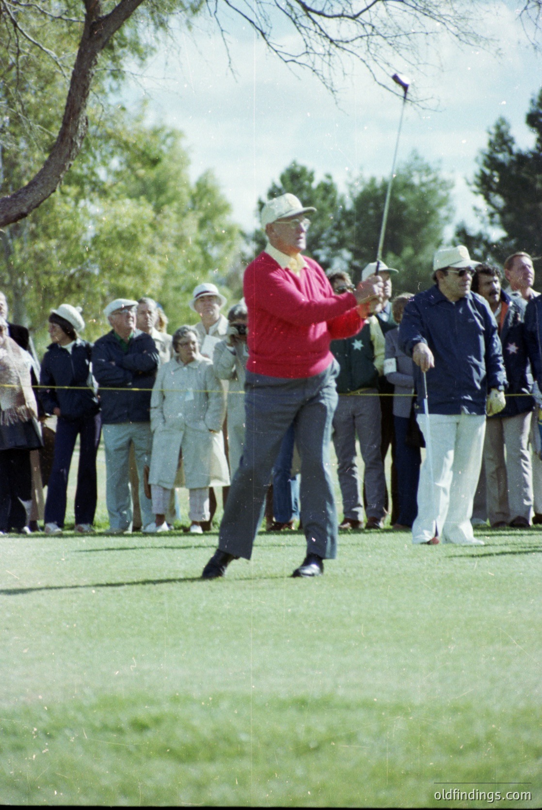 Mid-20th century golf scene: golfer in red sweater and white cap mid-swing on a lush green course, surrounded by spectators in vintage attire. Crowd stands behind ropes, suggesting a public or tournament event. Trees and clear sky indicate an outdoor setting, likely a sunny day. Classic sportswear and attire hint at 1950s–1960s era.