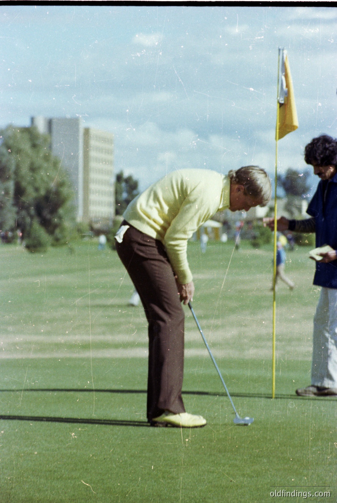 1970s golf scene: Male golfer in vintage sweater and trousers executes a putt on a lush green course. Urban high-rises and spectators in background suggest a city course. Classic mid-century golf attire and equipment highlight retro sports culture.