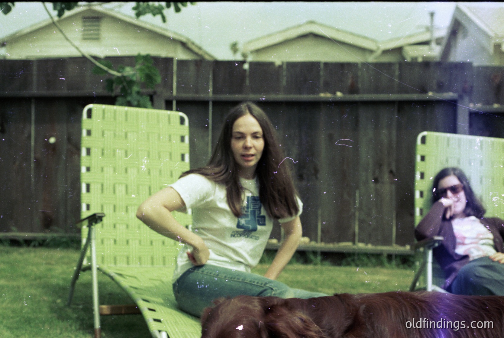 Vintage 1970s backyard scene: woman in long-sleeve graphic tee and bell-bottoms poses on a lime-green plastic lounge chair, with a dog resting at her feet. Blurred second person in sunglasses sits on a matching chair. Residential brick house with a flat roof and wooden fence in background.