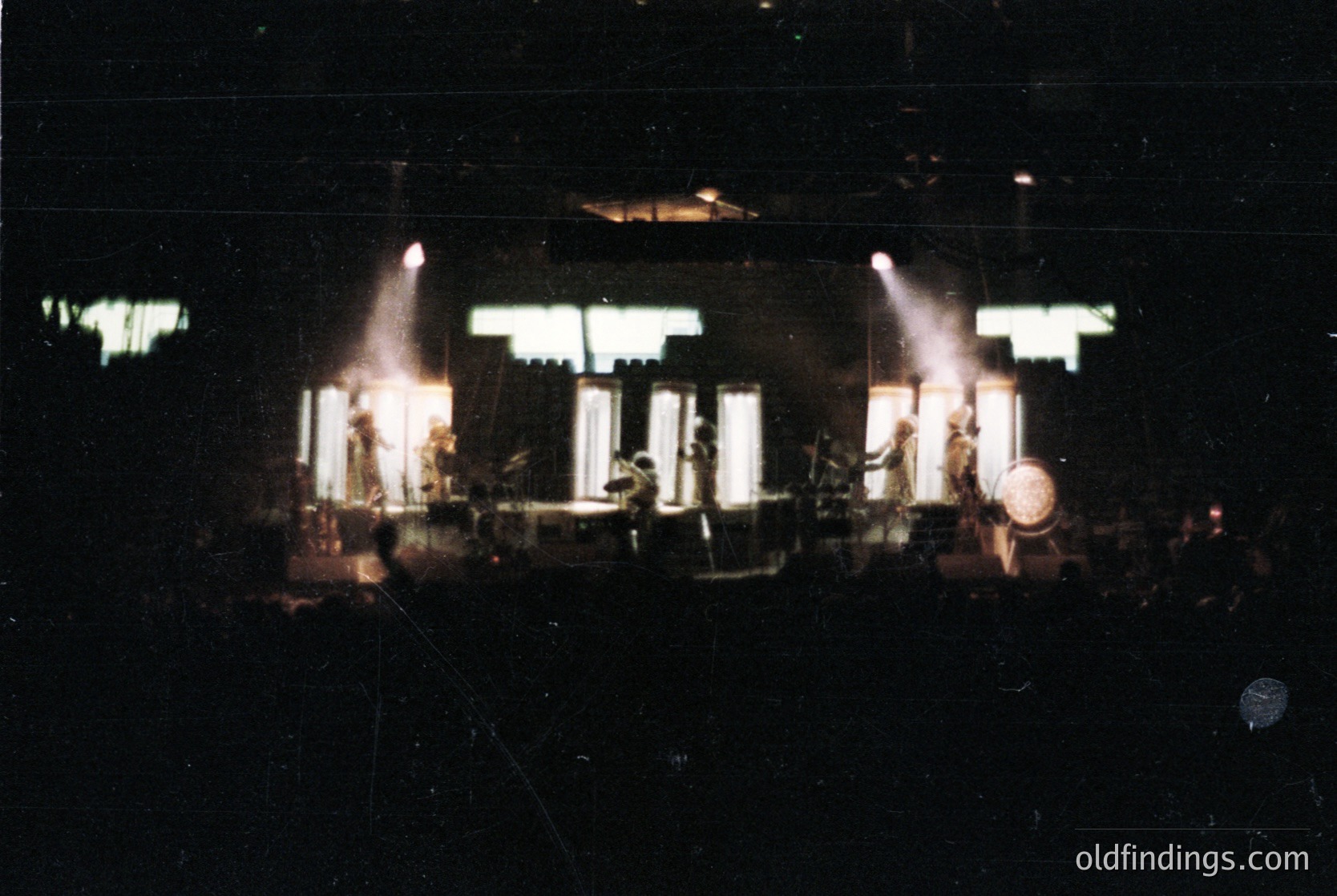 Vintage concert stage bathed in spotlight beams, featuring a drum kit and keyboardist silhouetted against dim lighting. Stage design includes vertical panel backdrops with geometric cutouts. Likely 1970s–1980s live music venue.