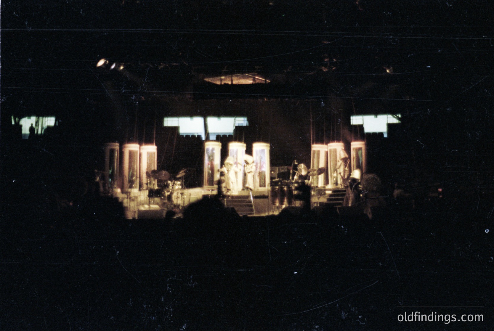 Vintage concert stage setup with spotlighted performers on raised platforms. Visible instruments include drums, keyboards, and microphones. Stage lighting features circular fixtures and vertical beams. Likely mid-20th century live music venue.