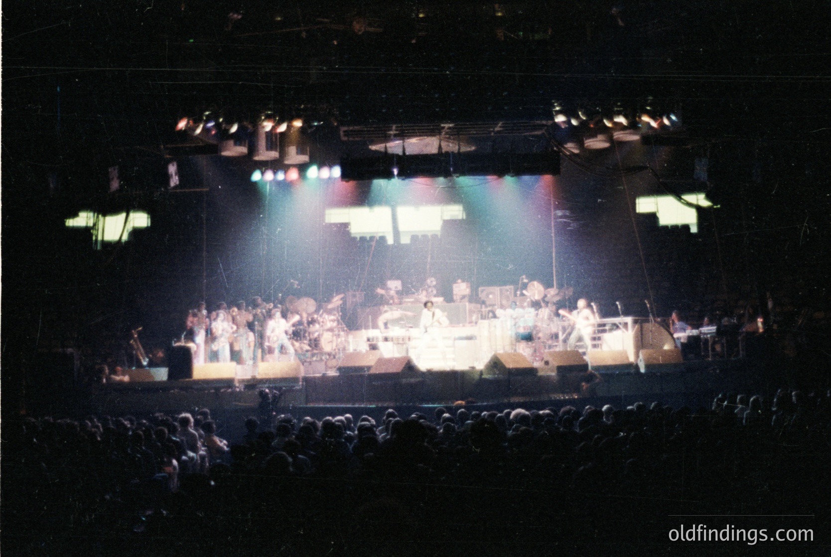 Vintage concert stage featuring a large band performing under colorful stage lights, likely 1970s–1980s. Drummer, keyboardists, and guitarists visible; audience silhouetted in foreground. Industrial lighting and geometric stage backdrop suggest a rock/metal venue.