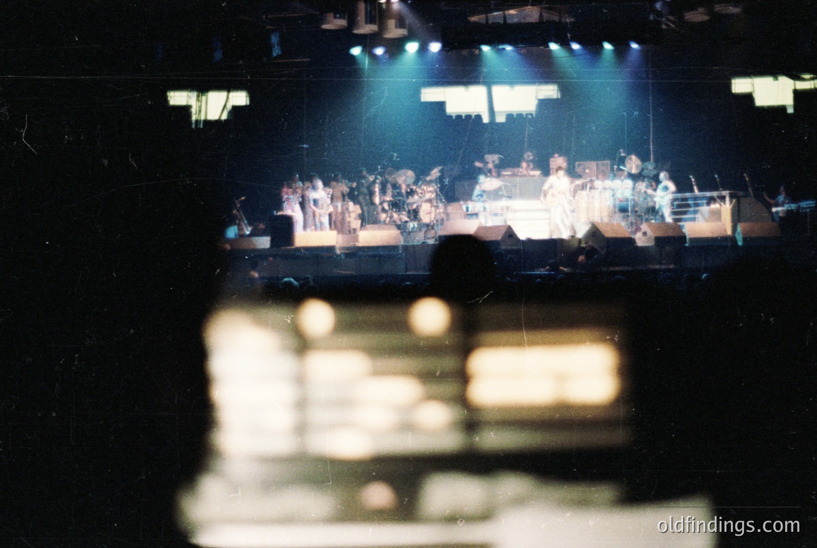 Vintage concert stage with band performing under blue stage lighting. Visible instruments include drums, keyboards, and guitars. Crowd silhouetted in foreground. Likely 1970s–1980s rock/psychedelic era.