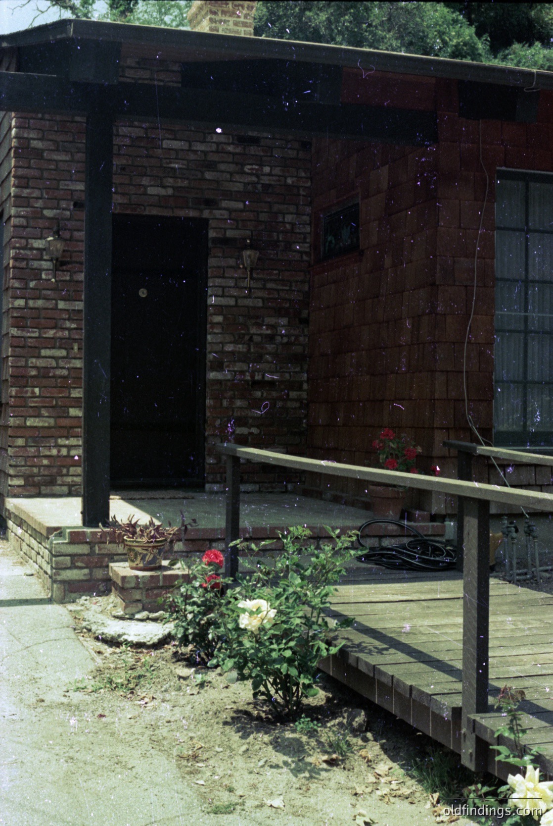 Mid-century brick entrance with concrete steps and wrought-iron railing, featuring a small landscaped garden with roses and potted plants. The dark wooden door contrasts with red brickwork, suggesting a residential or small commercial building from the 1950s–1970s.
