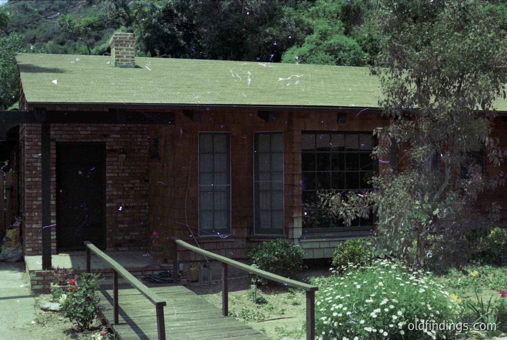 Mid-century modern brick home with green shingle roof, set in lush greenery. Brick foundation, large front windows, and a small porch with wooden railings. Likely 1960s-1970s suburban architecture.