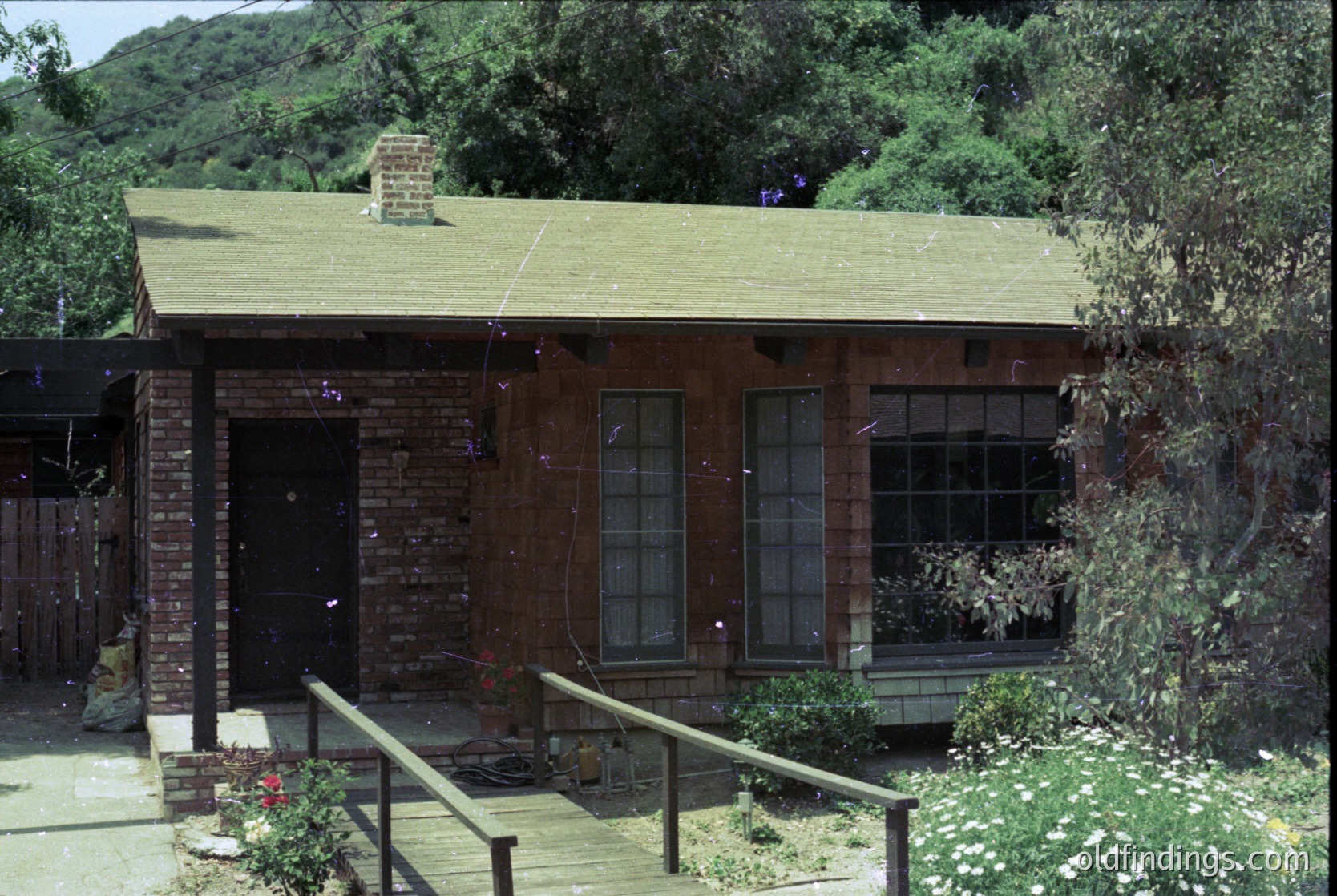 Mid-century brick cottage with gabled green roof, set in a lush, forested hillside. Brick chimney and large double-hung windows with flower boxes. Wooden deck with railing leads to entrance.