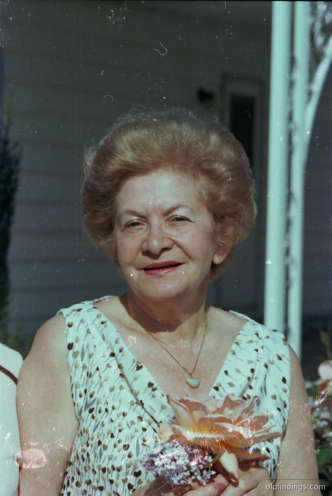 Vintage portrait of an elderly woman in a floral-patterned blouse, holding a bouquet of flowers. Soft focus and slight film grain suggest mid-20th century (1950s–1970s). Indoor setting with blurred background, likely residential.