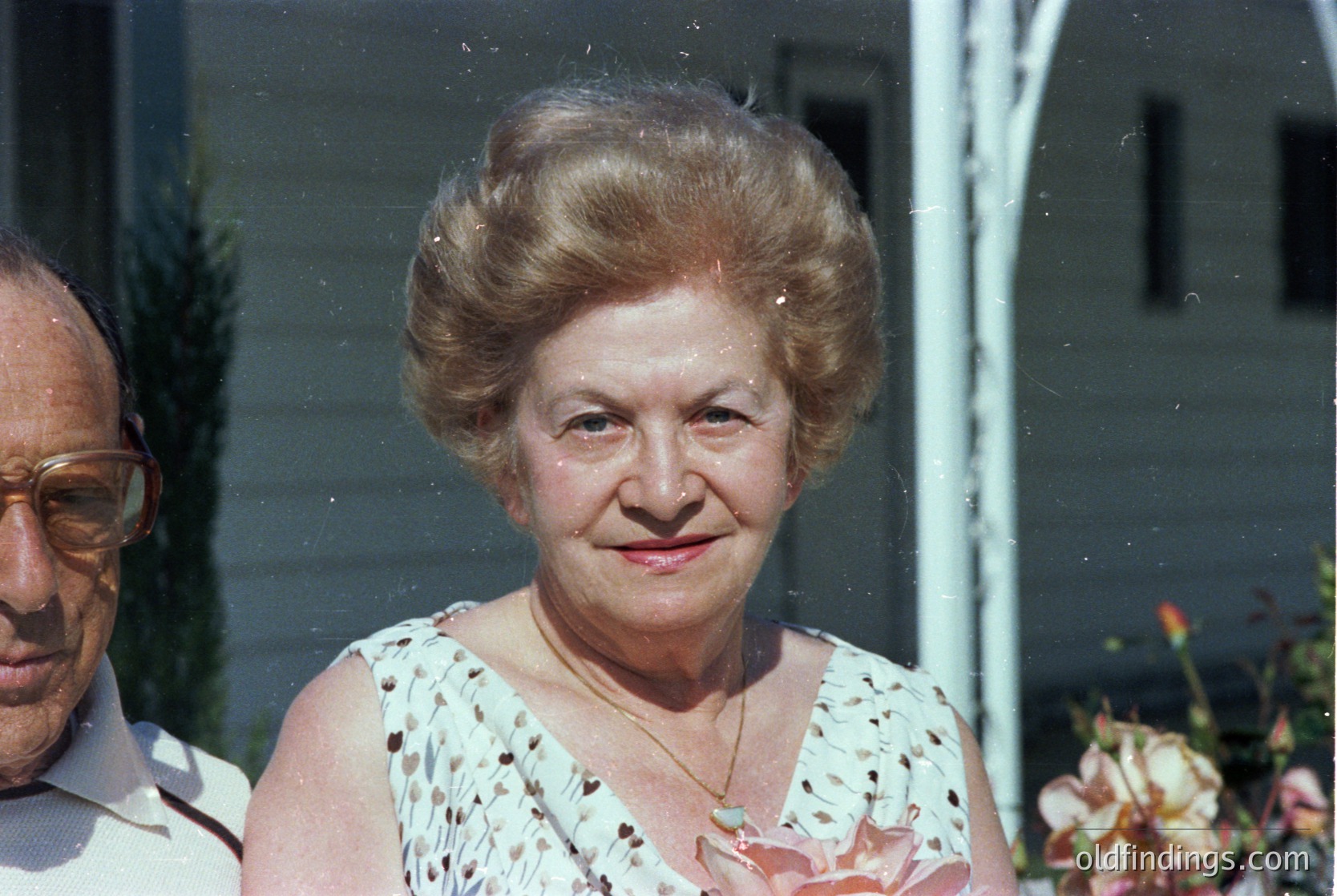 Vintage portrait of an elderly woman with voluminous 1970s-style updo, wearing a floral-patterned sleeveless blouse. Side profile shows classic mid-century hairstyle and subtle smile. Blurred background suggests indoor setting with light-colored walls and greenery.