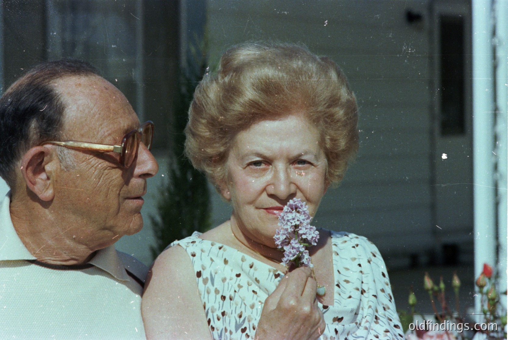 Mid-20th century portrait of an elderly couple, likely 1960s–1970s. Woman in floral-patterned blouse holds lilac blossoms; man wears round sunglasses and a light-colored shirt. Indoor setting with blurred greenery and building exterior. Candid, warm-toned snapshot captures quiet intimacy.