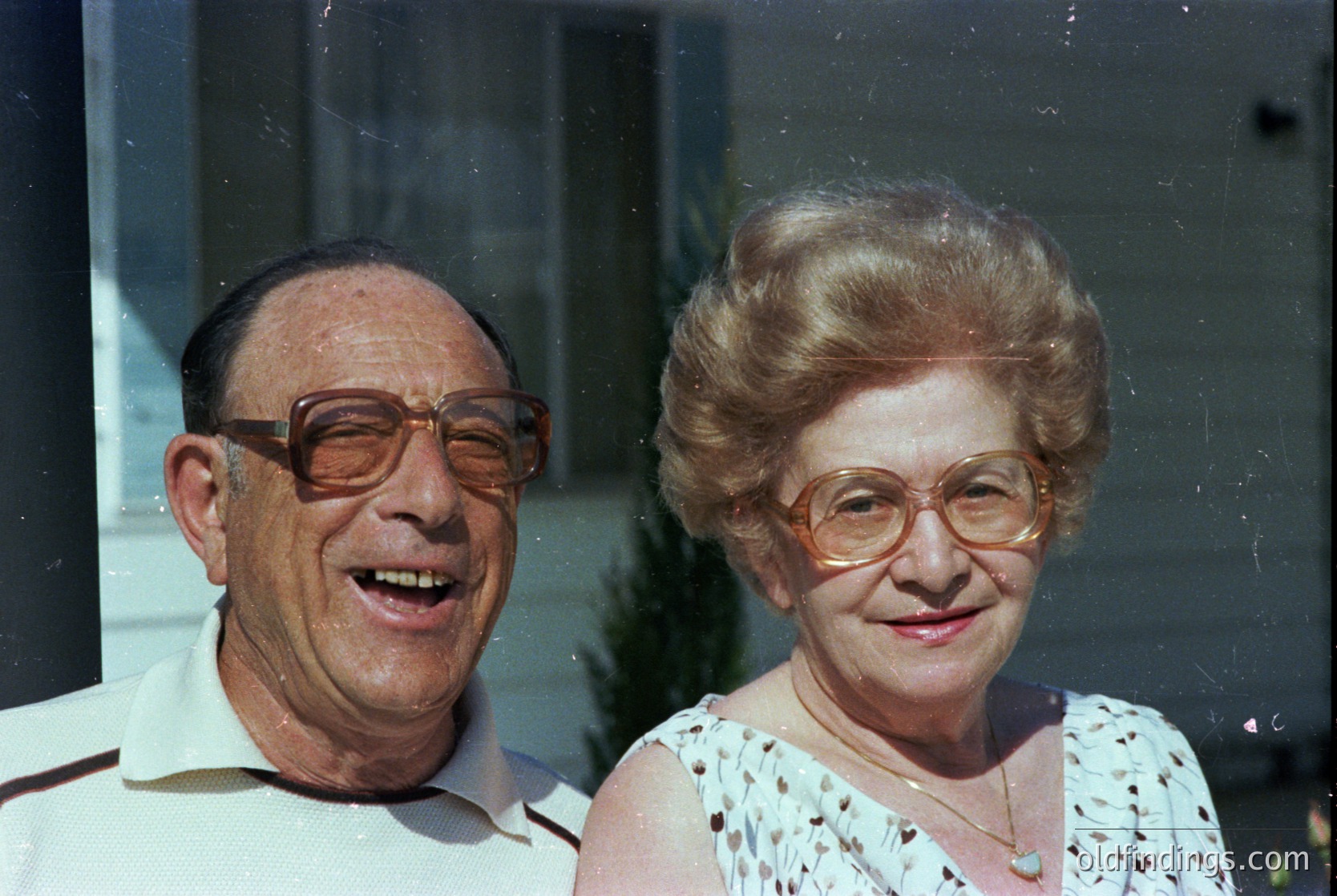 Retro portrait of an elderly couple posing indoors, likely mid-20th century (1960s–1980s). Man wears round, wire-framed glasses and a light-colored button-up; woman has short, voluminous hair and round glasses with a floral-patterned blouse. Soft lighting and blurred background suggest a candid, home setting.