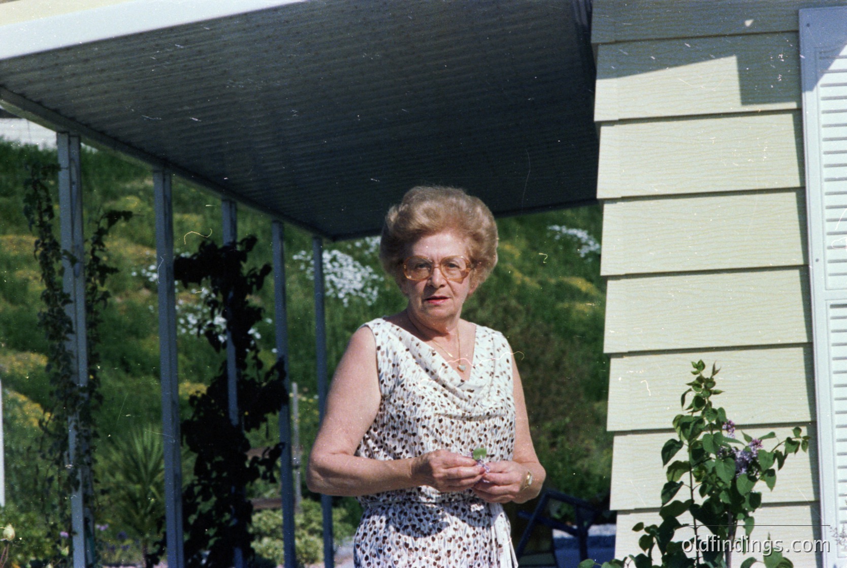 Mid-century woman in a floral dress poses under a covered patio, holding a small object. Light-colored wooden house with vertical siding and metal railings. Lush greenery and flowering plants in background. Likely 1960s–1970s suburban or rural setting.