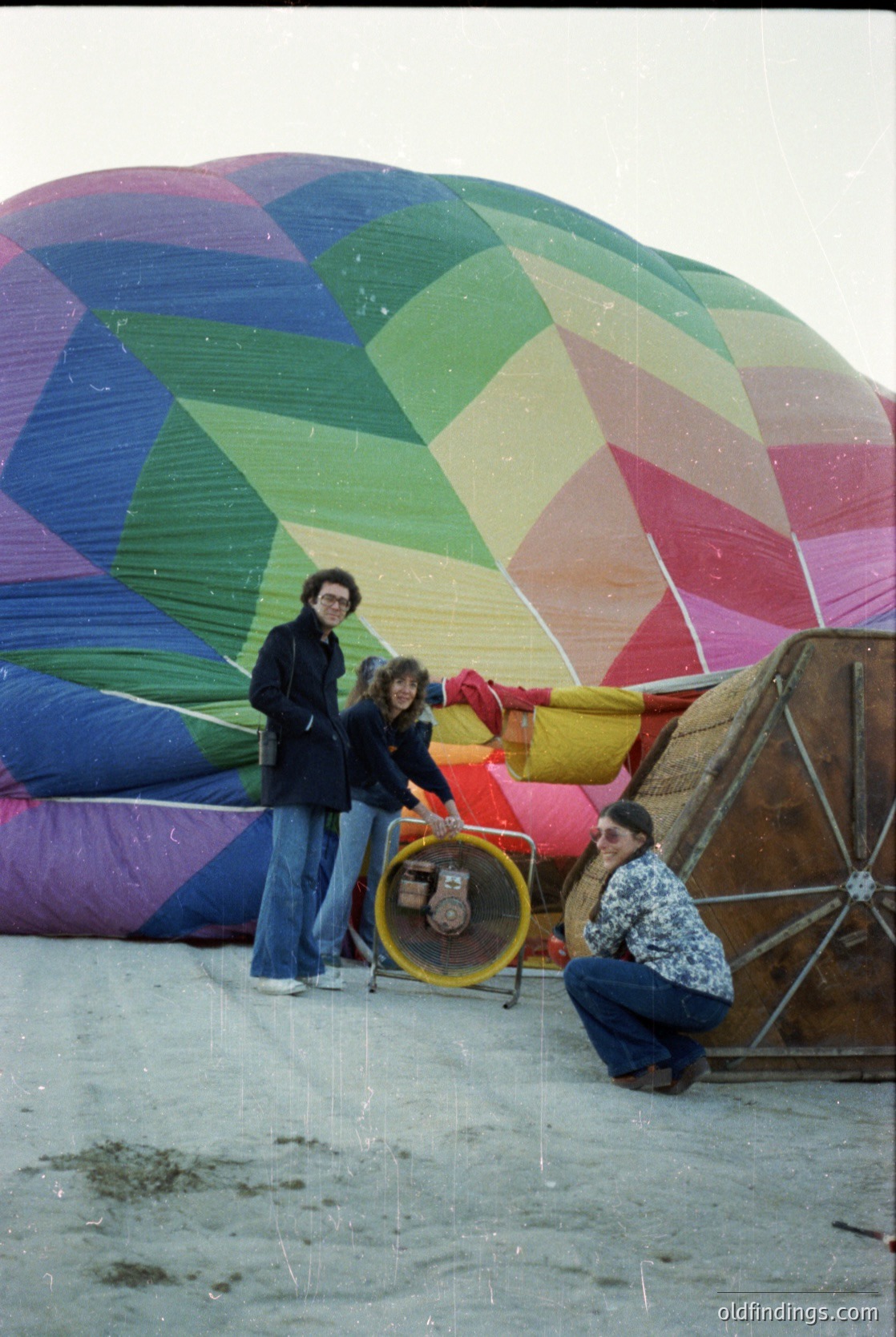 Vibrant 1970s-style hot air balloon with geometric, rainbow-colored panels being inflated by three people on sandy ground. Two standing, one kneeling near a yellow fan. Casual 70s fashion: wide-leg pants, patterned blouses, and layered jackets. Likely a beach or open-air event.