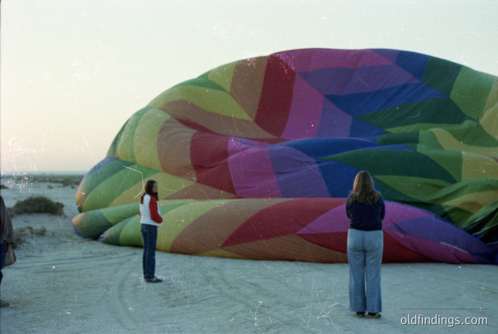 Two individuals stand on a wet beach, inspecting a large, geometric-patterned hot air balloon with vibrant stripes in green, red, and purple. The scene suggests a coastal setting with shallow water and sandy shore. Clothing indicates a casual, outdoor activity from the late 20th century.