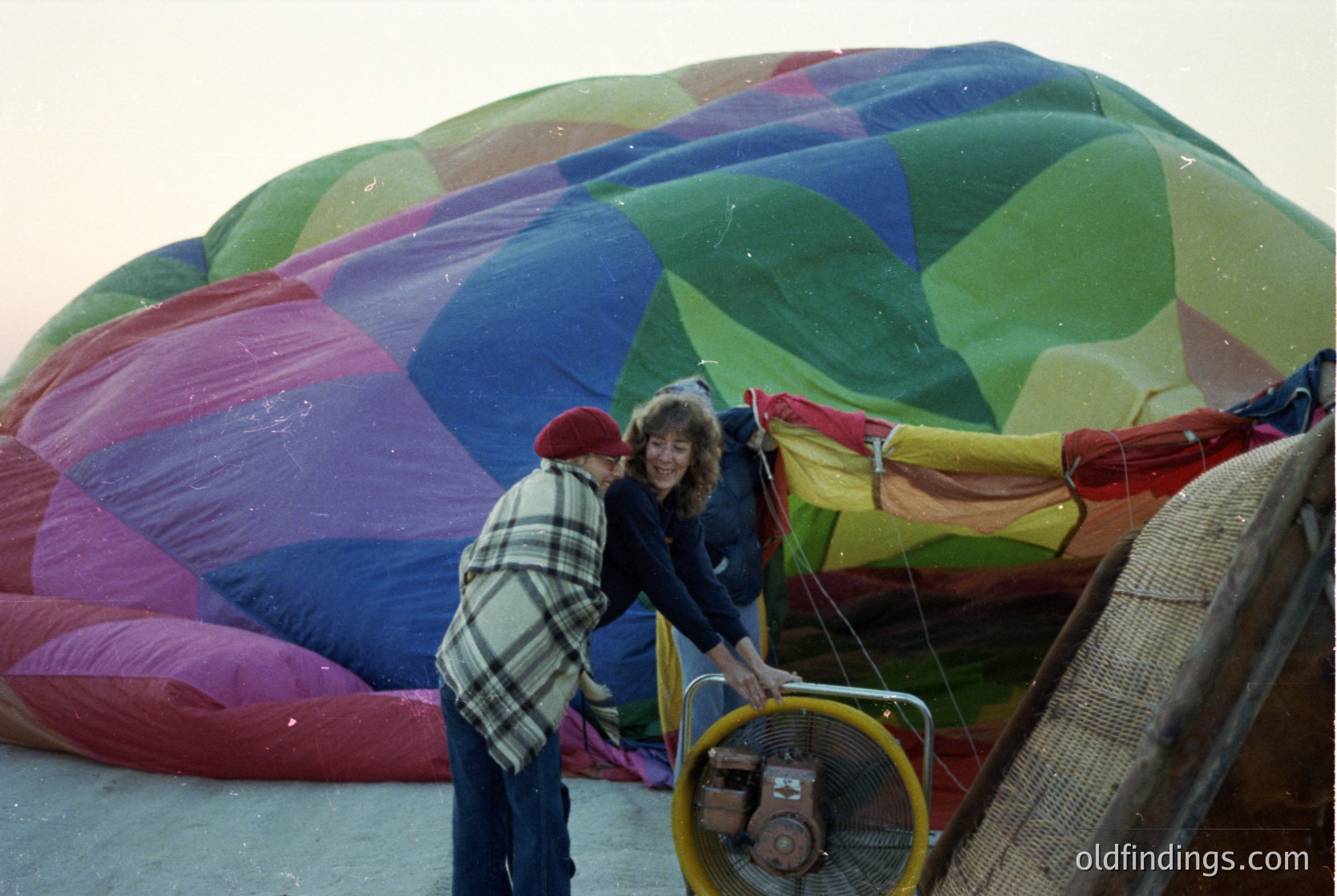 Vibrant 1970s hot air balloon in deflated state, with two people preparing for launch. The balloon’s fabric features bold geometric patterns in red, green, blue, and yellow. One person wears a red knit cap and plaid shirt, the other a dark jacket. A yellow fan and basket are visible, indicating ground operations.