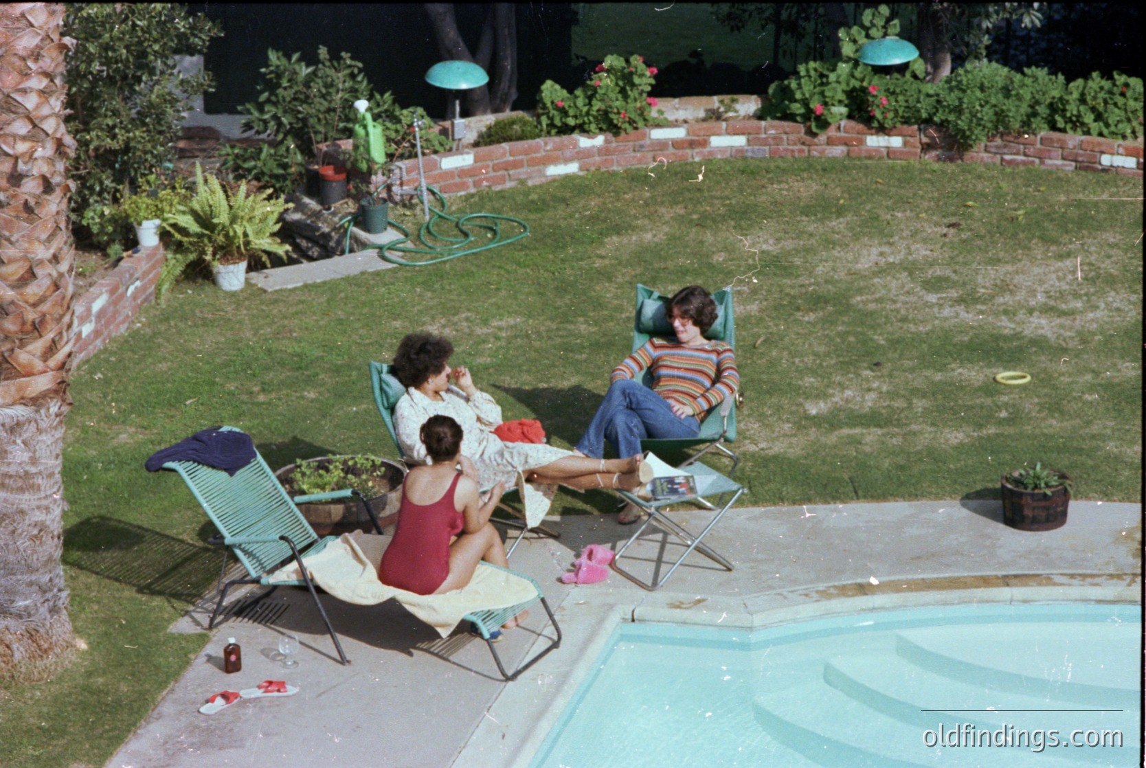 Vintage backyard pool scene with three adults lounging on green metal chairs, 1970s-80s suburban style. Concrete pool with shallow edge, brick planters, and teal umbrellas. Casual summer attire—striped shirts, shorts, and dresses. Side table holds a bottle cap and small bag.