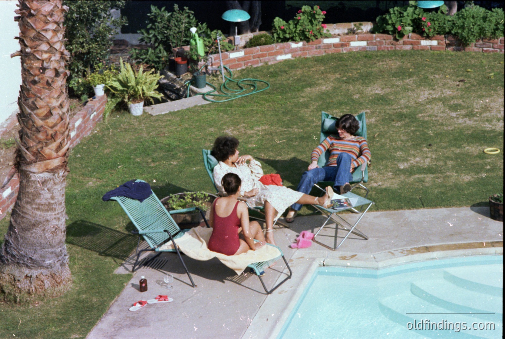 Aerial view of a mid-20th-century backyard pool scene. Three adults relax on vintage lounge chairs beside a rectangular pool, surrounded by manicured lawn, palm tree, and potted plants. Colorful towels and a toy hint at leisure.
