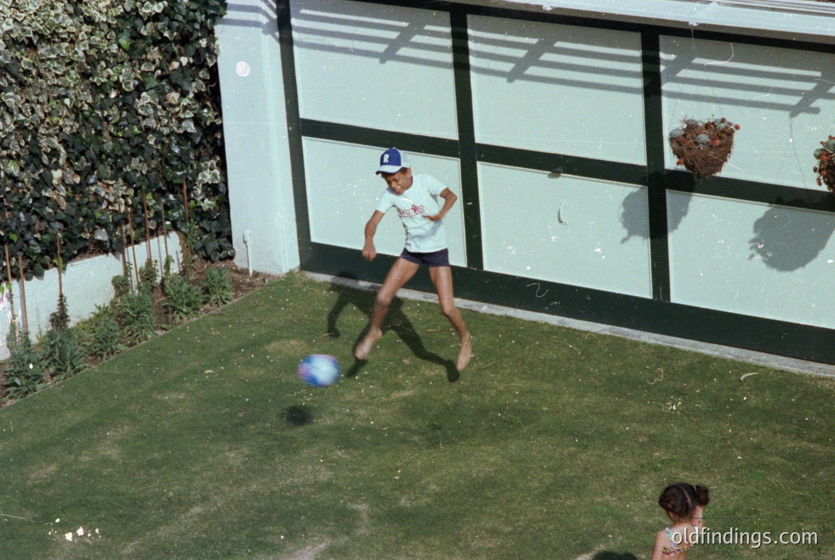 Mid-20th century suburban backyard scene: boy in white sports shirt and blue cap kicks a blue ball toward a garage door. Lush green lawn, potted plants, and a child seated on grass edge. Residential architecture reflects 1960s-70s style.