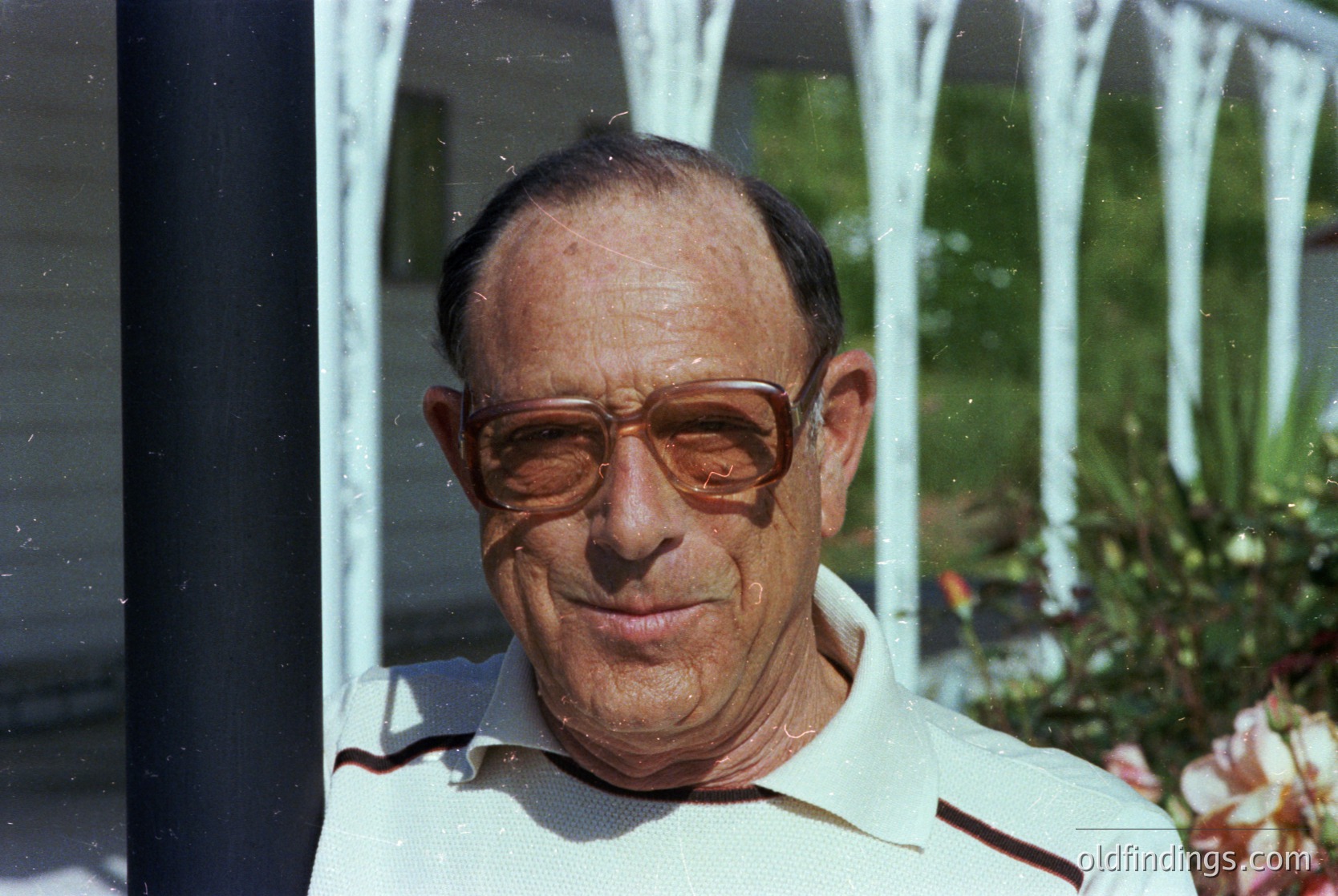 Portrait of an elderly man in vintage round-framed glasses, wearing a white polo shirt with red stripes. Reflections of a fountain and greenery in the background suggest an outdoor setting, likely a garden or courtyard. Style and lighting suggest mid-20th century (1960s–1980s).