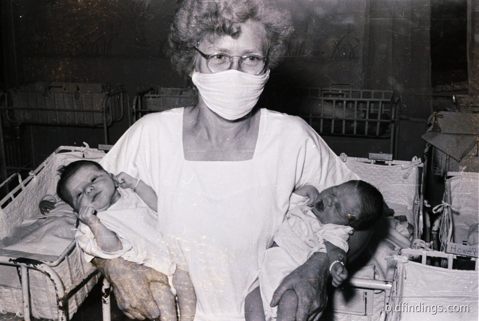 Mid-20th century hospital nursery scene: female nurse in white uniform and mask cradles two newborns in metal cribs, likely –. Industrial-era medical setting with visible medical equipment and signage (). Evokes themes of maternal care and early pediatric healthcare ().