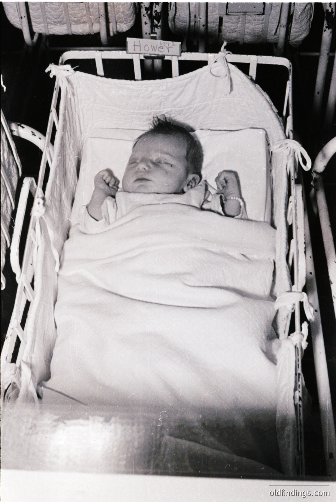 Vintage black-and-white photo of an infant in a metal crib, name tag reading "Howey." Soft cloth diapers and blankets wrapped around the child. Mid-20th century domestic setting, likely 1940s–1960s.