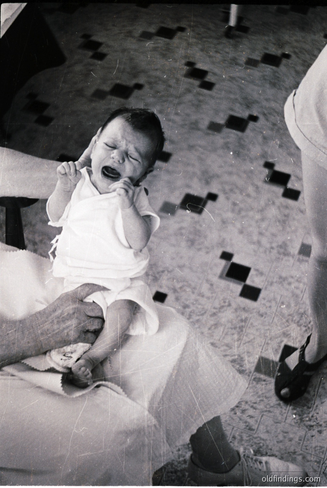Vintage black-and-white photo of an infant crying while being held by an adult in a clinical setting, likely a hospital or clinic. Checkered floor tiles and medical equipment (stethoscope, chair) suggest a 1950s–1970s healthcare environment. Candid, intimate moment captures early pediatric care.