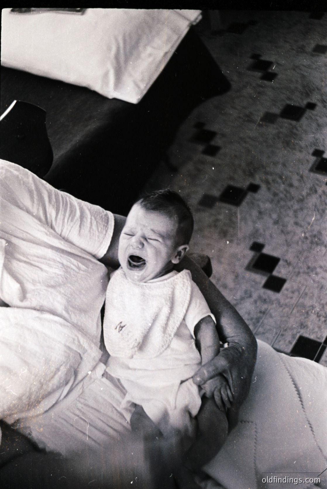 Black-and-white candid of an infant crying on a patterned rug, held by an adult’s arm in a domestic setting. Mid-20th century clothing and furniture suggest a mid-century home.