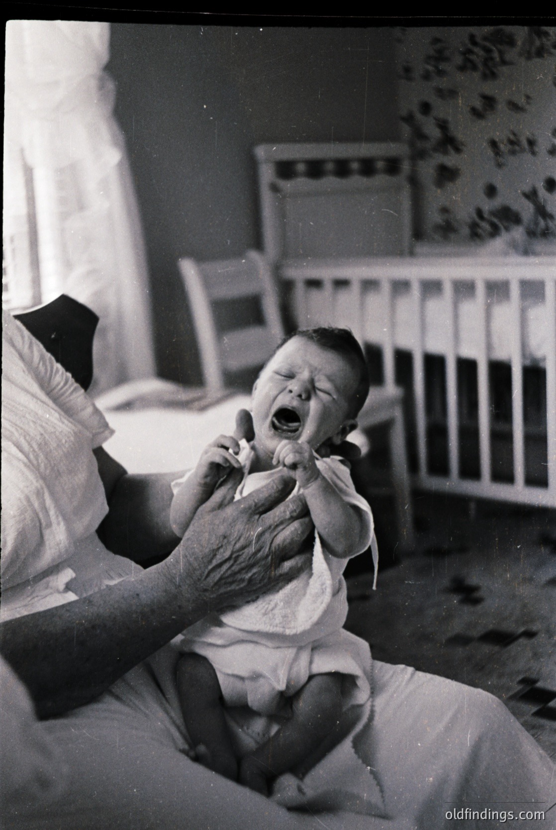 Mid-20th century indoor scene: elderly woman holding crying infant in a simple bedroom. Wooden crib with floral wallpaper in background. Classic 1950s-60s domestic setting.