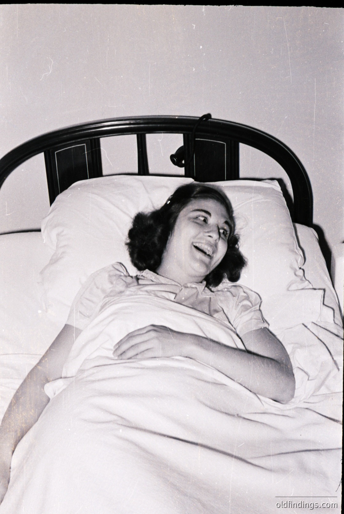 Mid-20th century hospital bed with metal frame, featuring a relaxed woman in a light hospital gown. Classic 1950s–1960s medical ward ambiance.