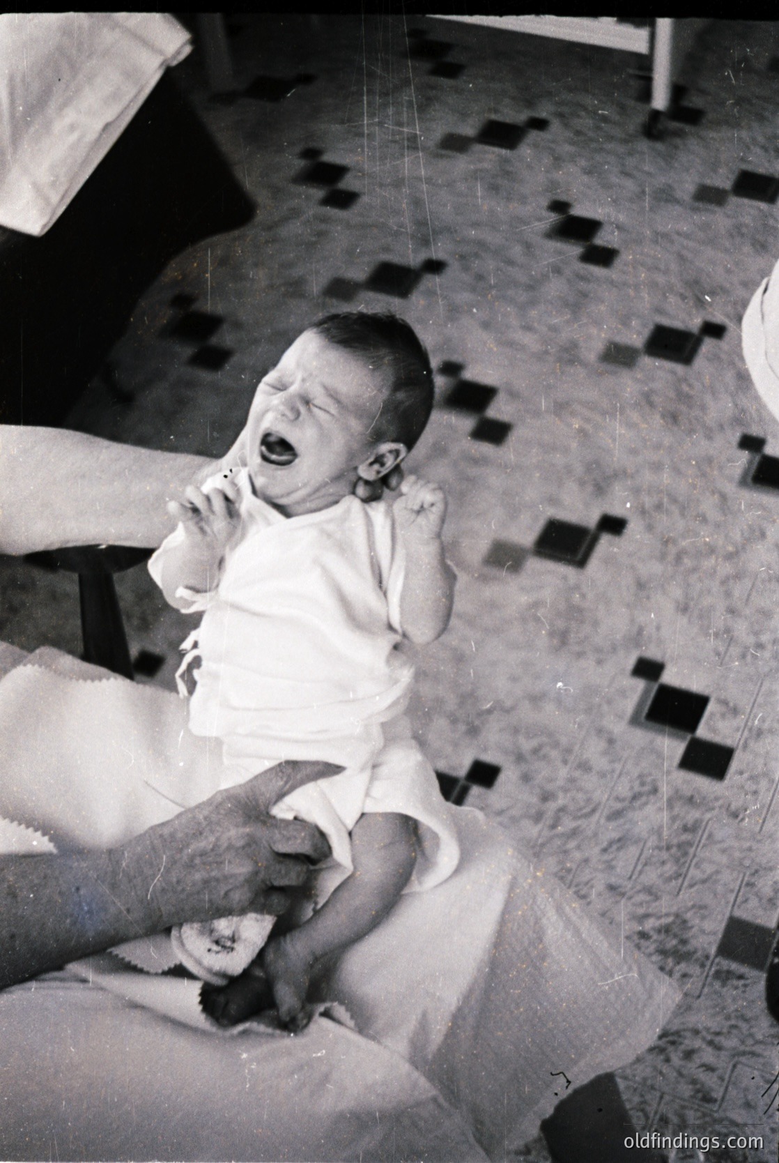 Mid-20th century black-and-white photo of an infant crying in a highchair, wearing a diaper and short-sleeve onesie. Checkered floor tiles and a partial view of a wooden chair frame suggest a domestic setting. Likely or home interior.