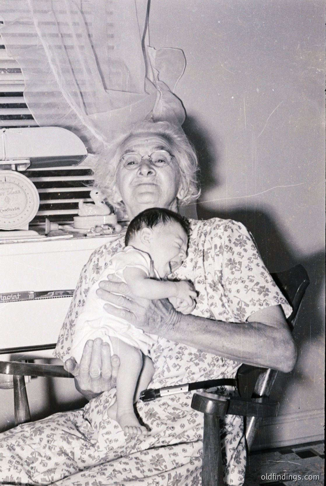 Mid-20th century black-and-white portrait of an elderly woman cradling a baby in a floral dress, seated on a wooden chair. Background shows vintage kitchen appliances, including a radio and a boxed item labeled "Vitamins." Warm, intimate domestic scene.