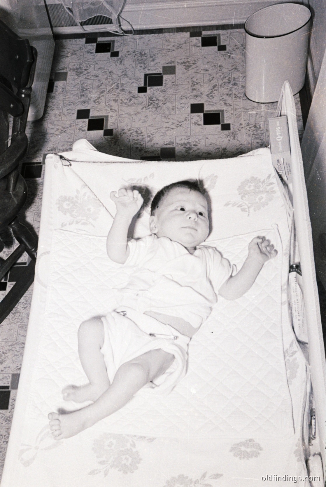 Vintage black-and-white photo of an infant lying on patterned bedding in a crib, mid-20th century. Checkered floor tiles and a metal bucket visible in background. Classic mid-century domestic interior.