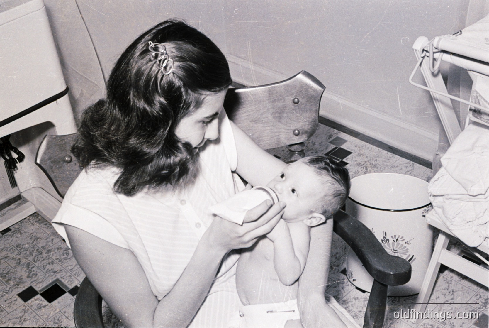 Nurse in 1950s-60s uniform bottle-feeding an infant in a clinical setting, likely a hospital or clinic. Metal feeding chair and vintage enamel basin visible.