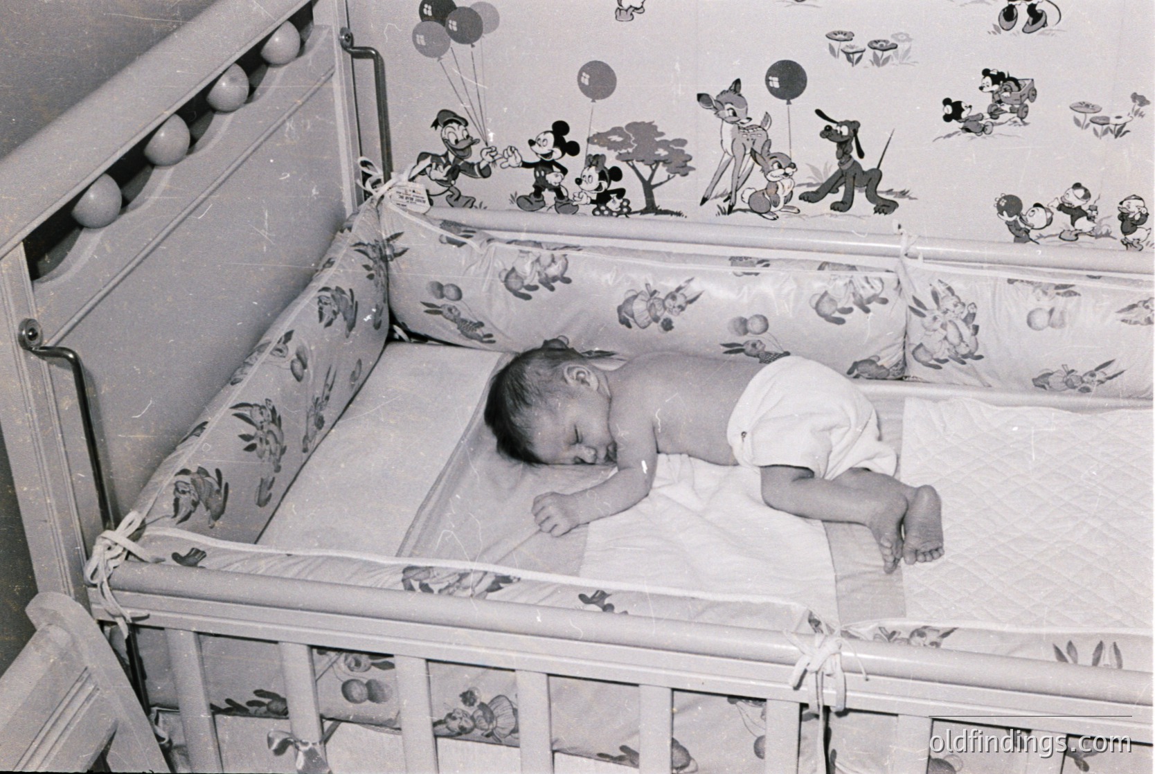 Mid-20th century baby crib with Mickey Mouse-themed decor, featuring cartoon characters and balloons. Infant sleeps peacefully in a classic wooden crib with floral-patterned railings. Likely 1950s–1960s, Western pop culture era.