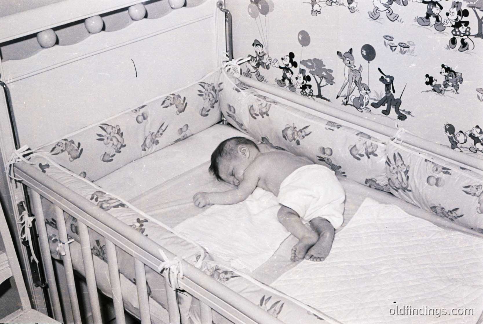 Vintage black-and-white photo of an infant sleeping in a crib adorned with 1960s cartoon-themed bedding (Mickey Mouse, Donald Duck). Wooden slat design with rounded corners. Soft focus and nostalgic mid-century aesthetic.