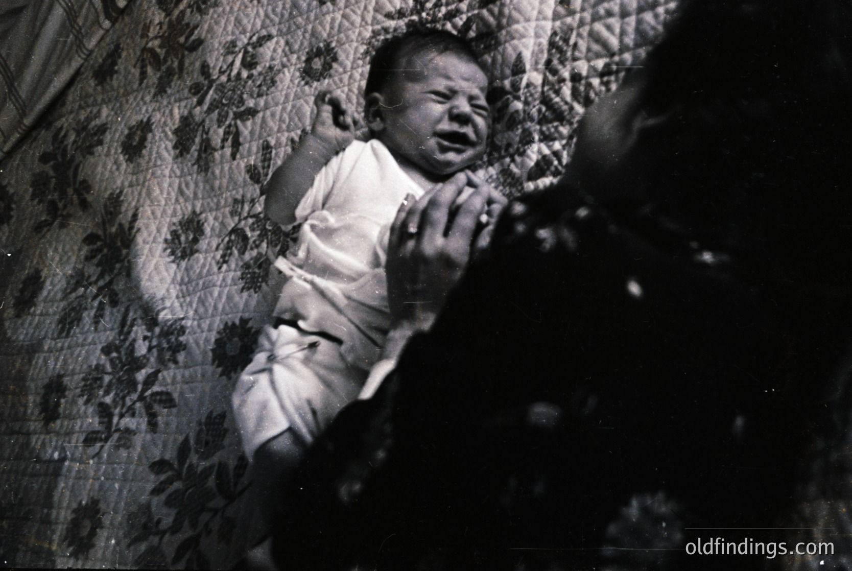 Black-and-white portrait of an infant crying in a crib, draped in floral-patterned bedding. Mid-20th century domestic interior, likely 1950s–1960s.