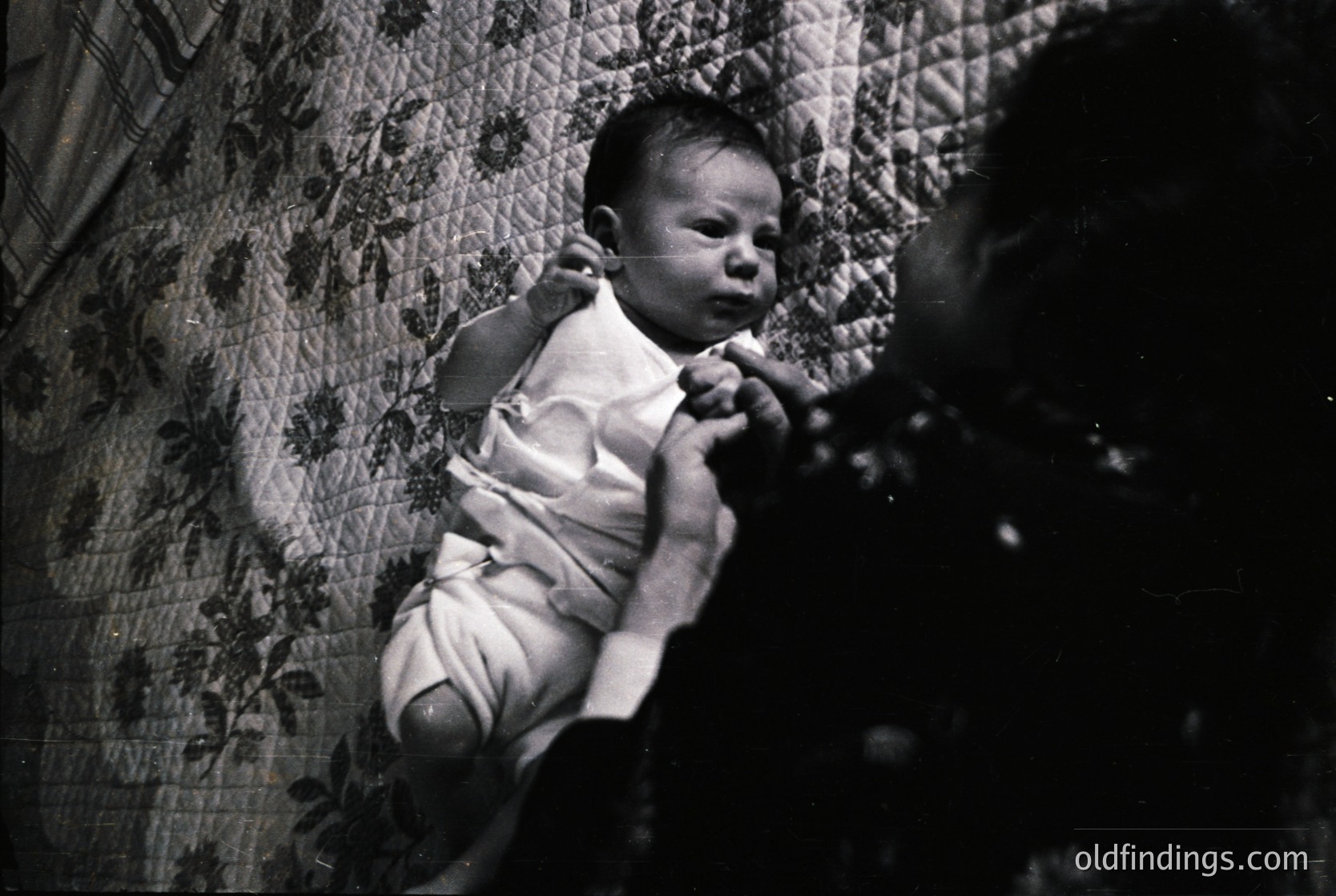 Vintage black-and-white photo of an infant in a diaper, suspended mid-air by an unseen adult’s hands. Floral-patterned wallpaper and dark, patterned curtains frame the scene. Likely mid-20th century (1940s–1960s), evoking nostalgia for early parenting practices.