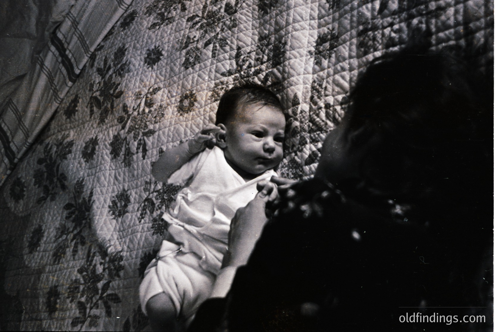 Black-and-white candid of an infant in a patterned quilted bed, mid-cry, with a blurred adult’s hand offering comfort. Mid-20th century domestic interior suggests warmth and intimacy. Ideal for historical research on parenting or vintage photography.