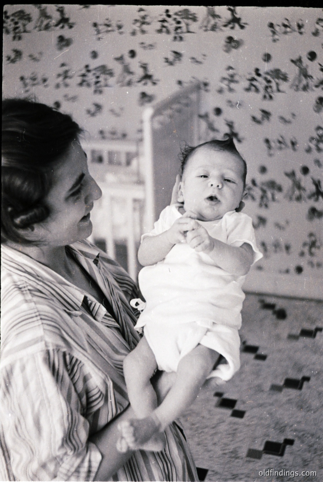 A woman cradles an infant in mid-1960s indoor setting, likely a home or clinic. Floral wallpaper with geometric floor tiles and a framed poster in background.