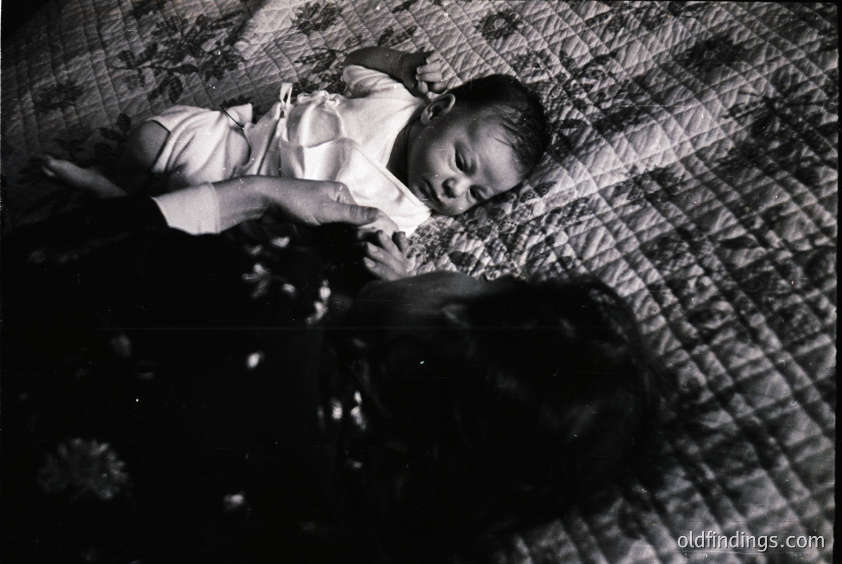 Mid-20th century black-and-white photo of an infant in a simple white dress, lying on a textured quilted blanket. The composition captures tender innocence with soft, diffused lighting. Style suggests vintage family photography, likely 1950s–1960s.